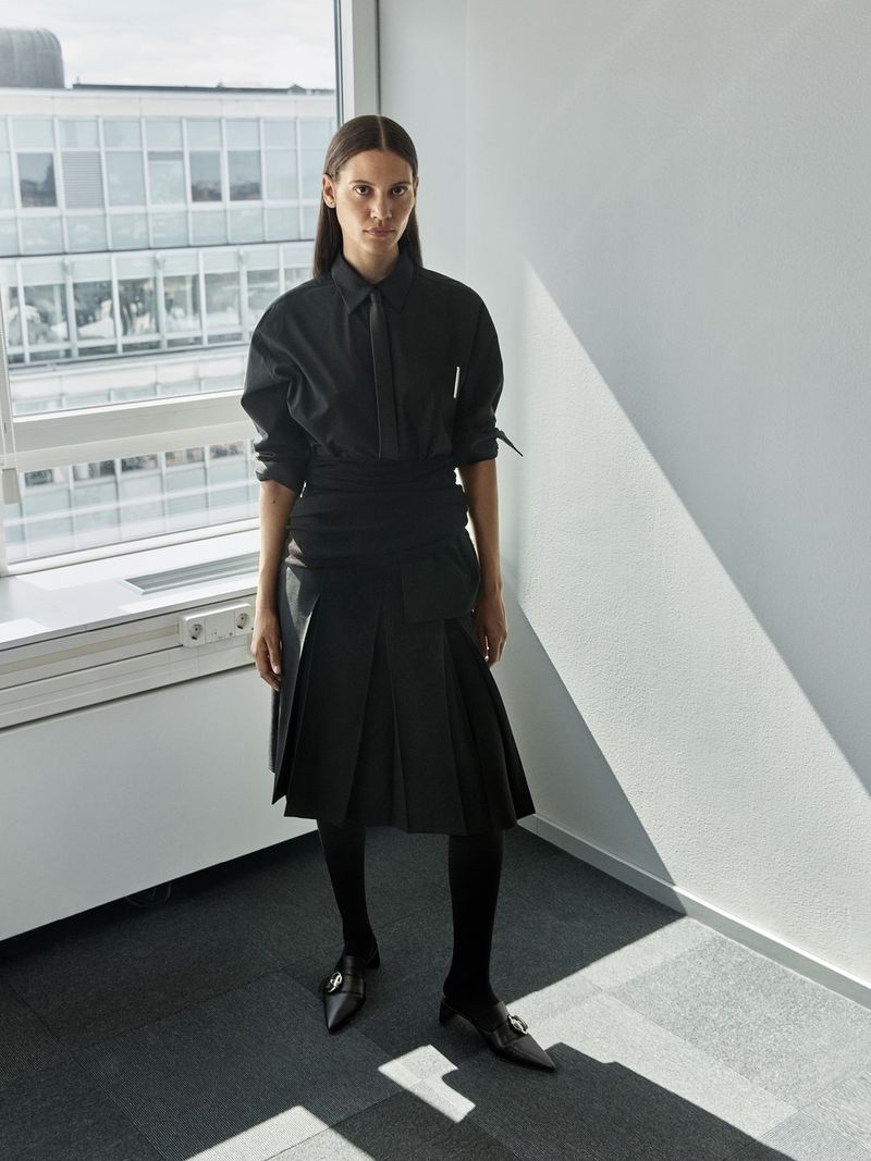 Woman in black button-down shirt and pleated skirt poses near window in a minimalist interior, with diffused natural light illuminating the scene.