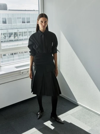 Woman in black button-down shirt and pleated skirt poses near window in a minimalist interior, with diffused natural light illuminating the scene.