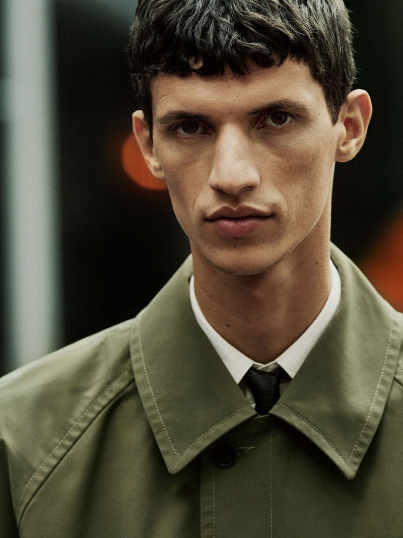 Close-up portrait of a young adult male wearing an olive-green overcoat, white collared shirt, and dark tie against a blurred backdrop.