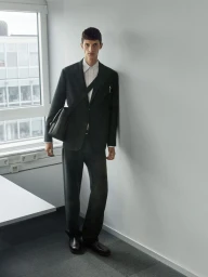 Model in a dark suit standing against an interior wall next to a large window carrying a black messenger bag