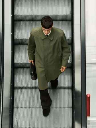 Man wearing an olive green coat and black trousers descending an escalator holding a briefcase
