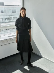 Model standing indoors by a large window wearing a black collared shirt, matching tie, and pleated skirt for Filippa K.