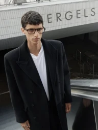 Model in a black coat and glasses standing near an escalator entrance for Filippa K Autumn/Winter 2025.