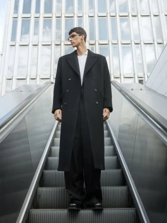 Man standing on an outdoor escalator wearing a long black coat against a modern building exterior.