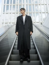 Man standing on an outdoor escalator wearing a long black coat against a modern building exterior.