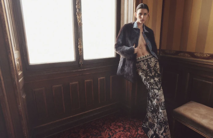 Model leaning against a tall window in a wood-paneled room, wearing a furry jacket over a denim shirt and patterned skirt.