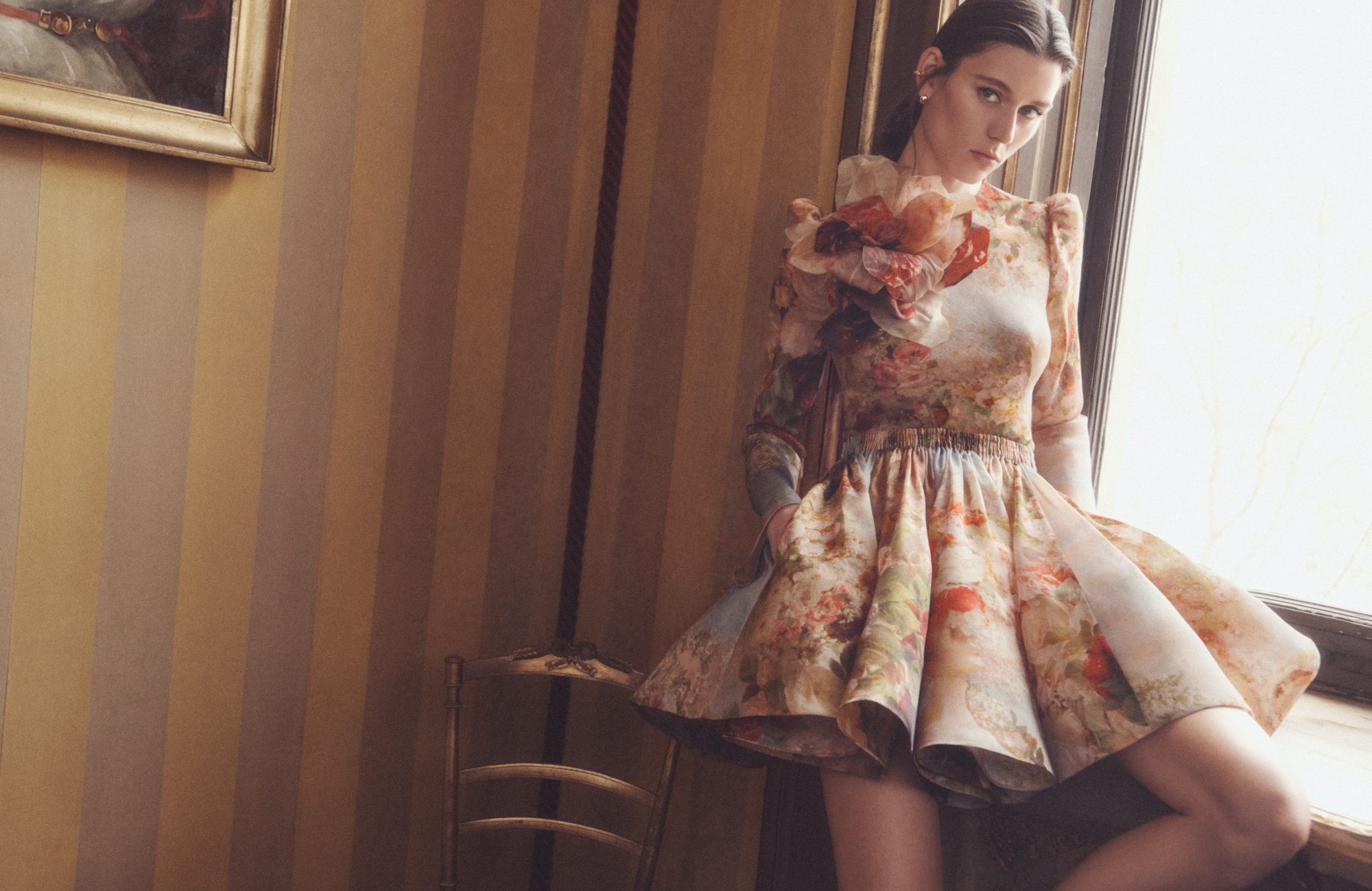 Indoor shot of a woman in floral dress, posing by a window. The dress features a ruffled neckline. Light-filled interior, painted walls.