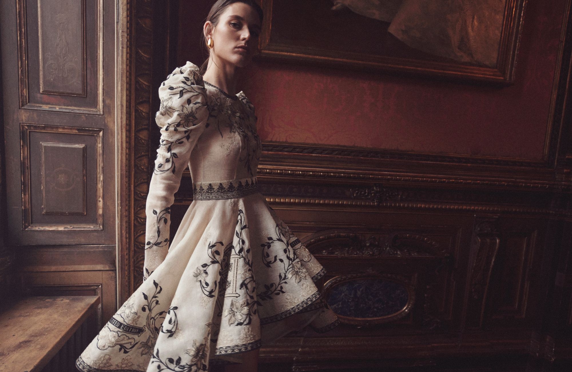 Elegant adult female in light floral dress poses in a vintage indoor setting with ornate details and warm, inviting lighting.