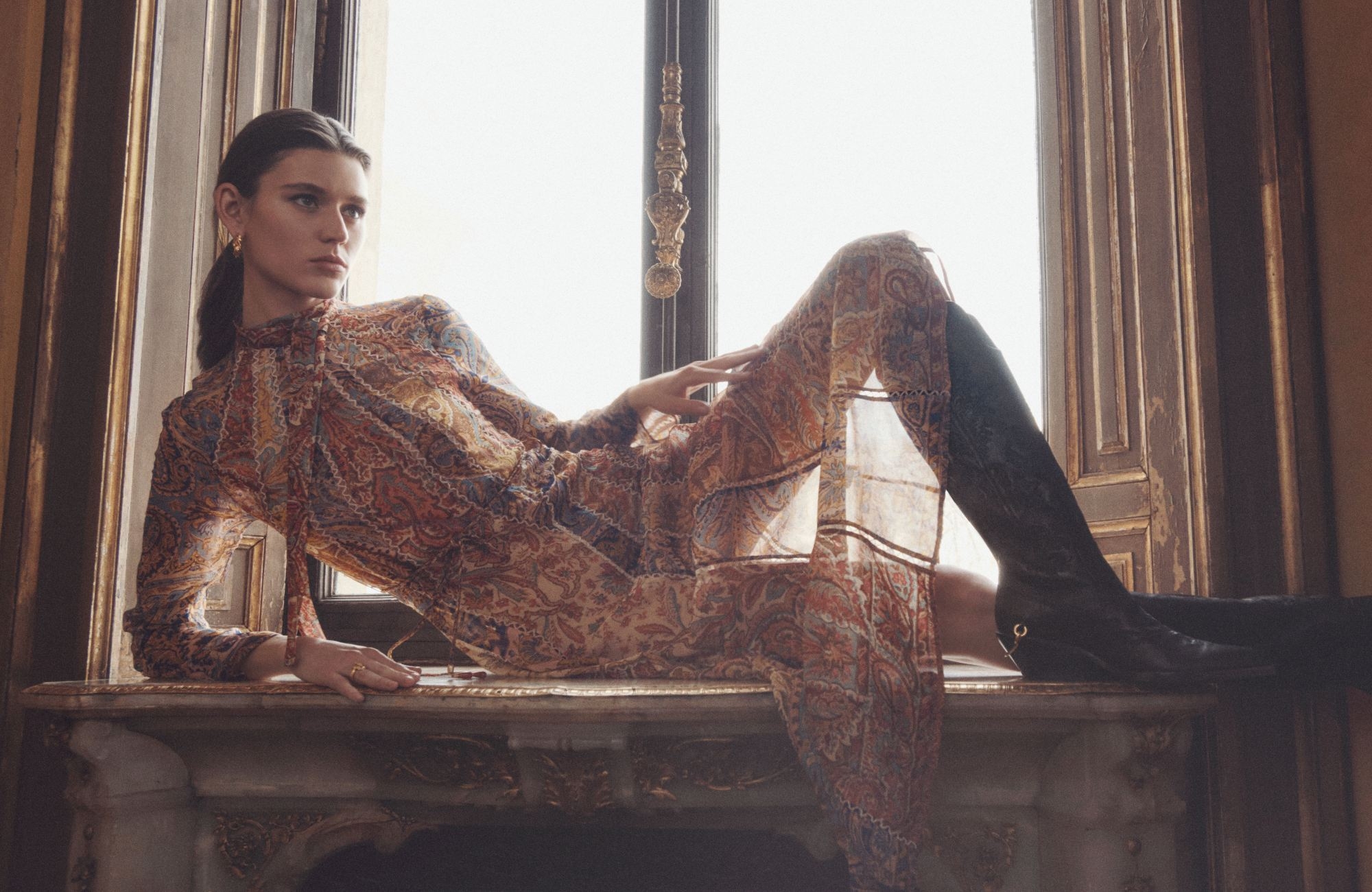 A woman in a patterned dress and black boots poses in front of a window, lit by bright daylight, inside a room with neutral walls.