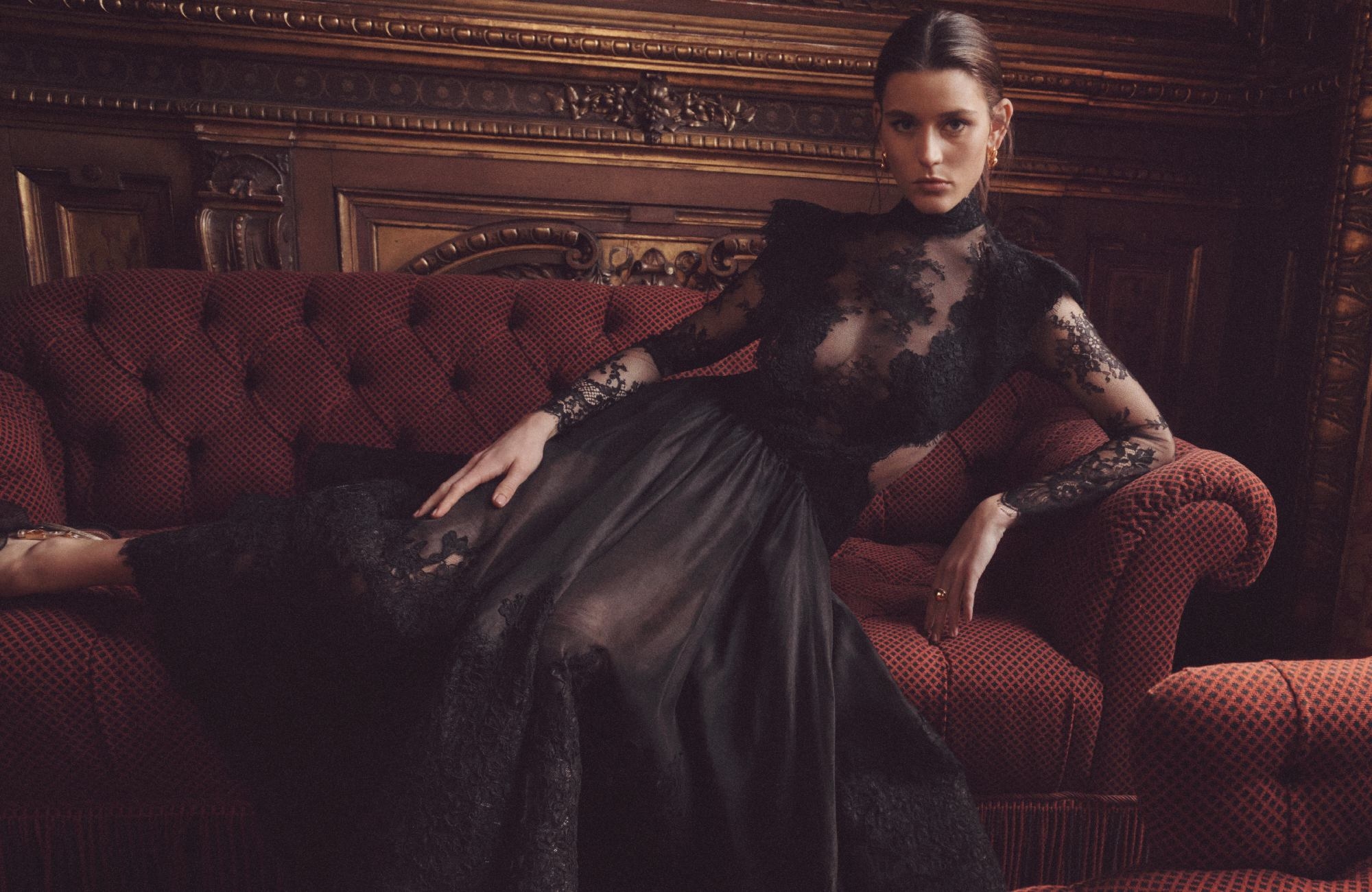 Adult woman in a dark, lacy, sheer dress poses on an antique red couch in an ornately decorated indoor setting with dark wood trim.