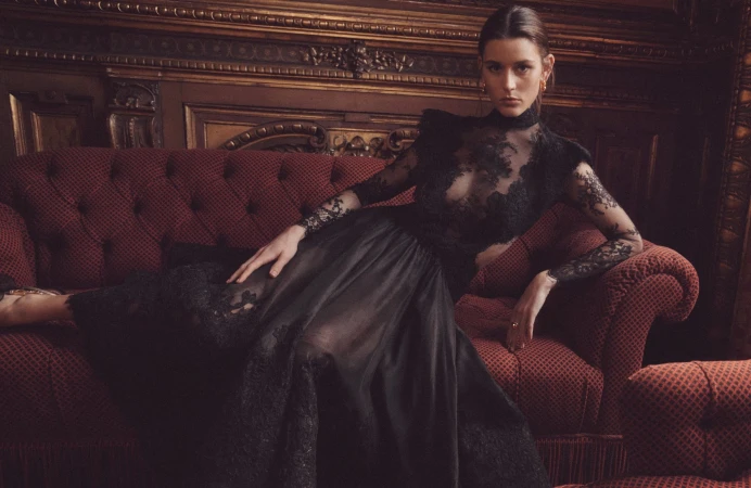 Adult woman in a dark, lacy, sheer dress poses on an antique red couch in an ornately decorated indoor setting with dark wood trim.