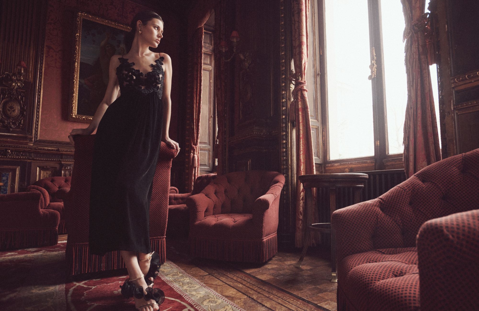 An elegant woman in a black evening gown stands in a lavishly decorated room with ornate details, plush armchairs, and a large window.