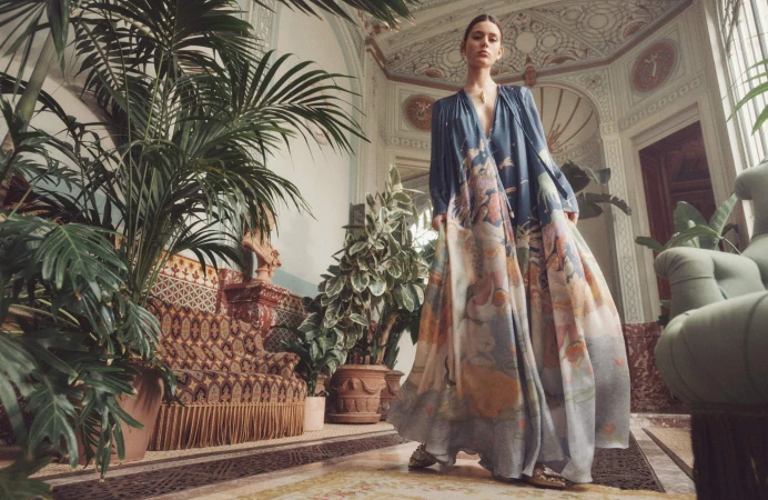 Model wearing a printed long dress standing in an ornate historic interior surrounded by potted palm plants