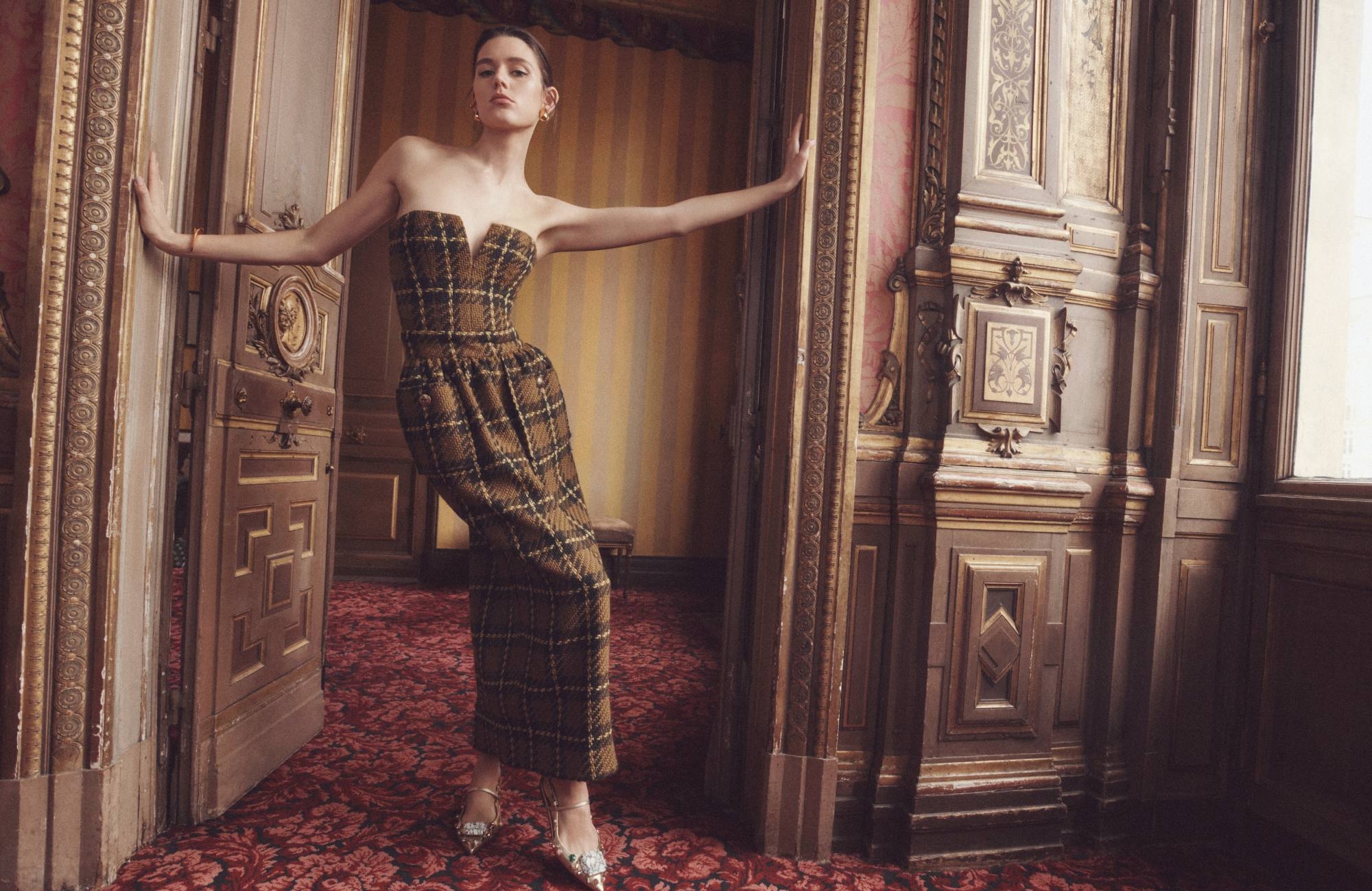 A woman poses in a plaid strapless dress, leaning against an ornate doorway, complemented by jeweled heels in an elegant interior setting.
