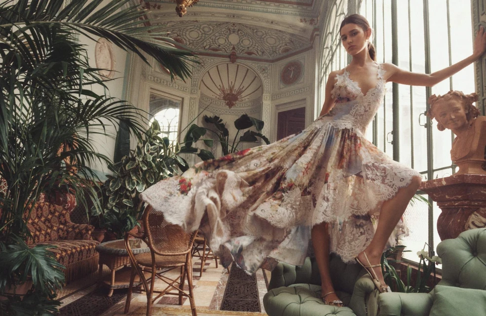 Fashion editorial shot of a young woman in a floral lace dress, leaning against a window in an ornate, plant-filled interior.
