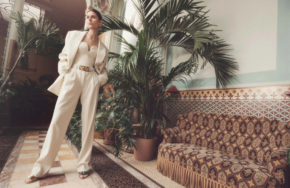 An adult female model poses indoors in a cream-colored suit, accessorized with a gold belt and black sandals, set against a backdrop of plants and patterned walls.