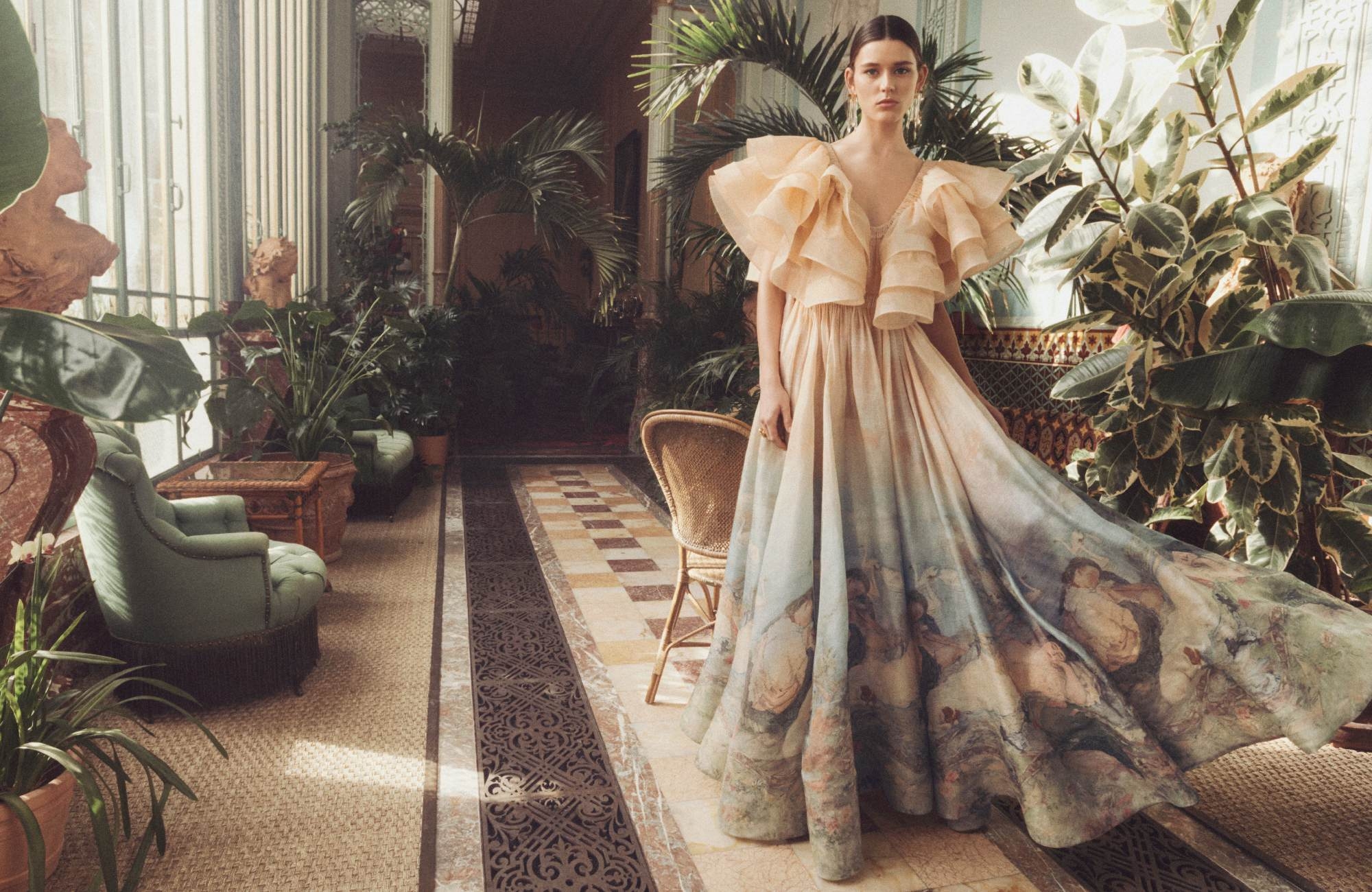 Woman in a pale ruffled gown poses in a sunroom filled with lush green plants and vintage furnishings, creating a lush and elegant scene.