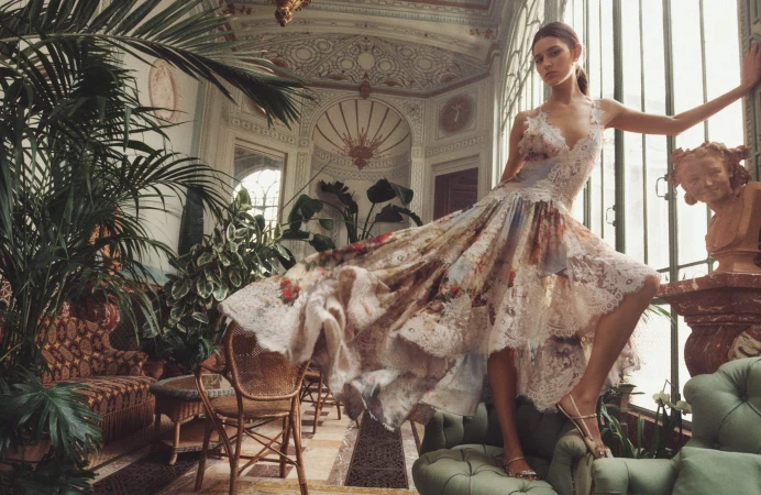 Model in a floral lace dress posing inside an ornate conservatory filled with potted plants and classical architecture.