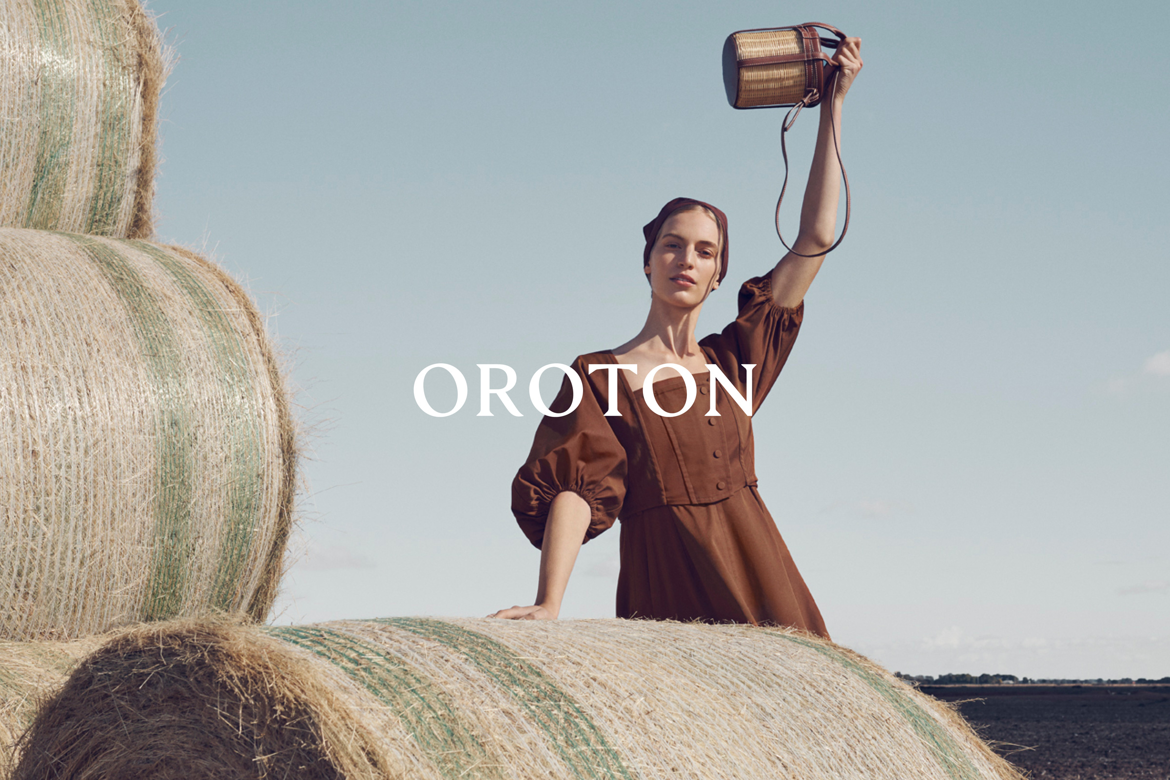 Woman in brown dress poses in a field with hay bales, holding up a woven purse against a blue sky, showcasing fashion and rural scenery.