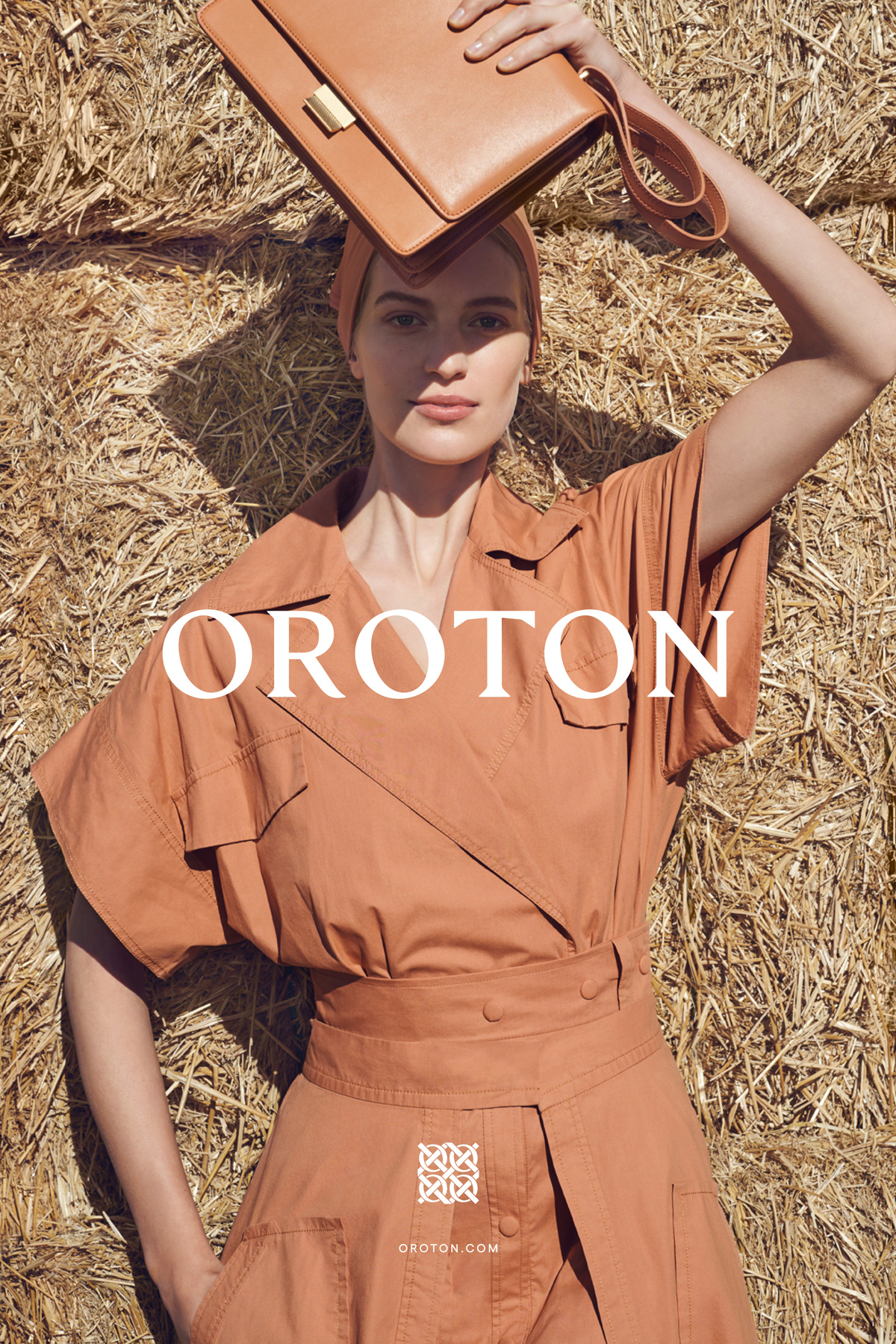 Fashion shot: Woman in earth-toned dress with handbag, lying on hay. Oroton brand, warm tones, sunny day. Fashion editorial advertisement.