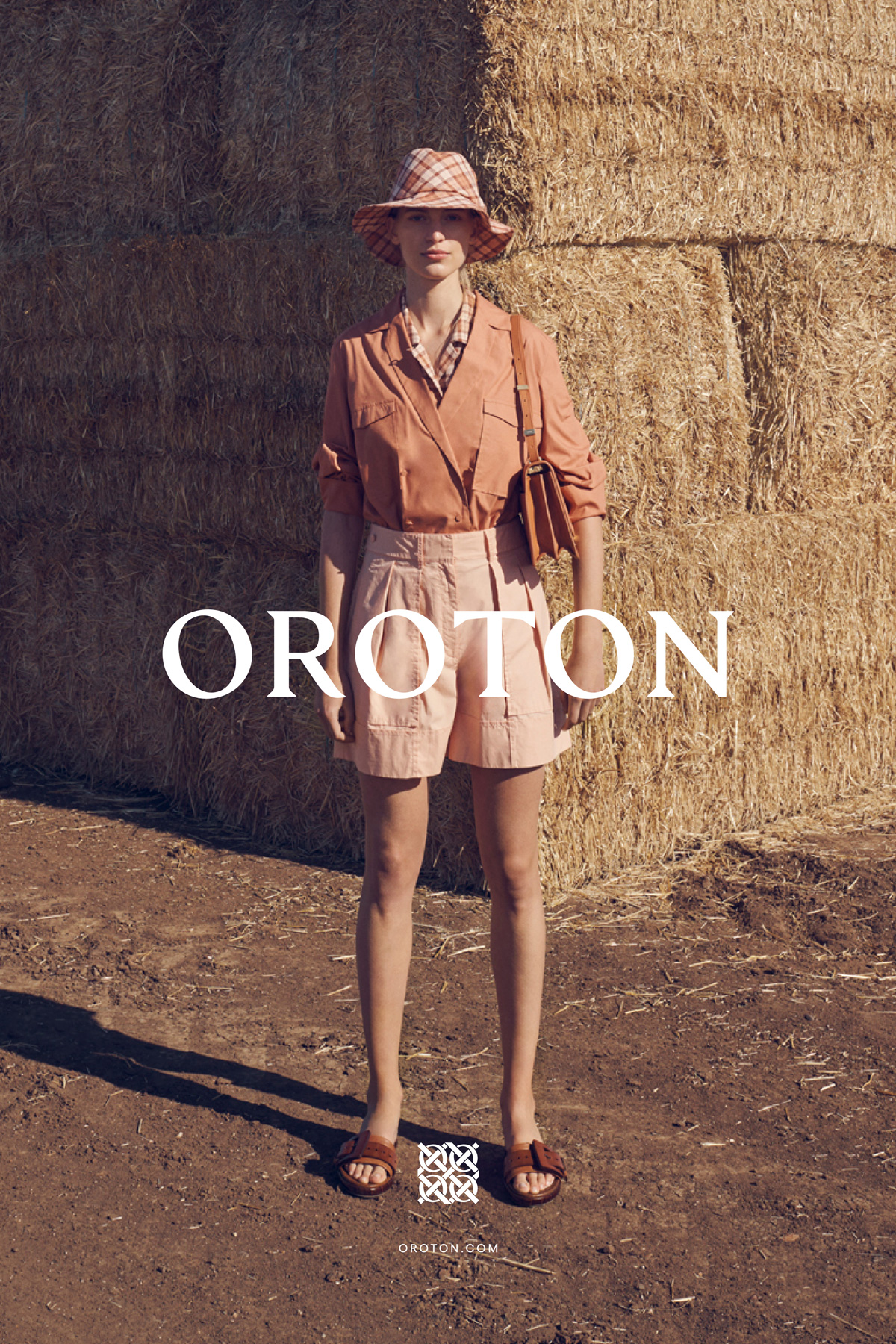 A woman in a matching shirt, shorts, and hat stands outdoors on a dirt surface in front of hay bales in a fashion advertisement.