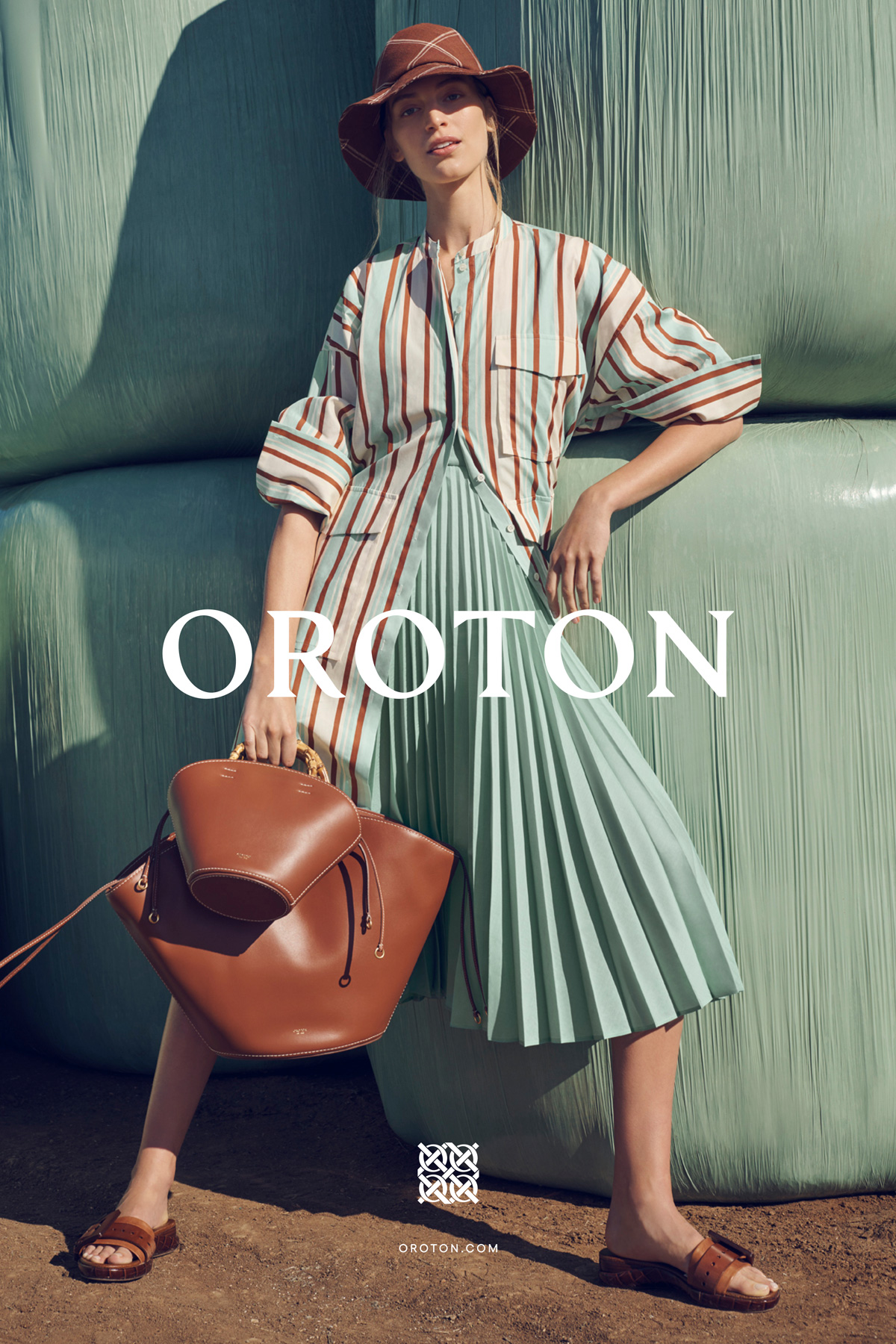 A woman in a brown hat, striped jacket, and pleated skirt poses with a brown handbag in front of stacked green hay bales.