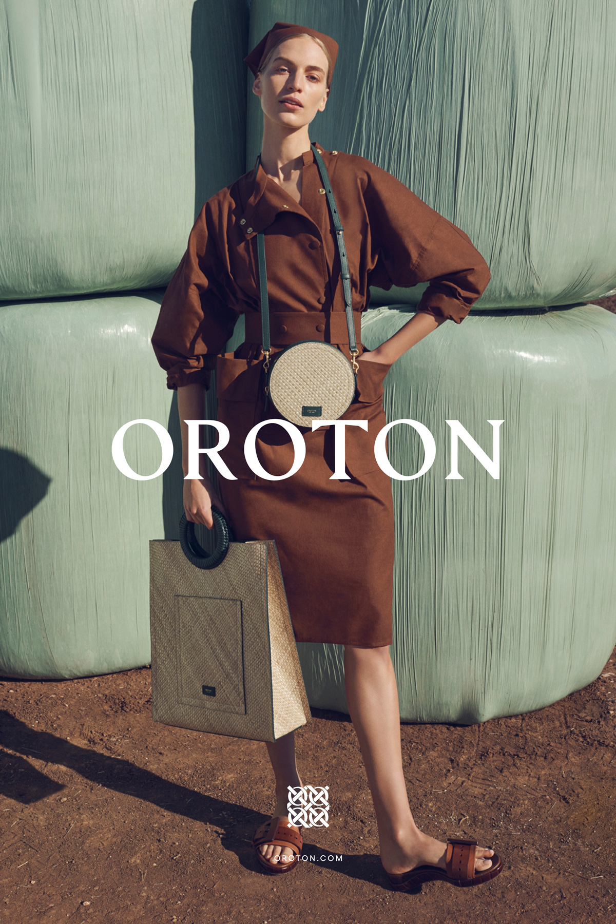 Fashionable woman in brown dress holds tote bag in front of stacked hay bales, wearing cross-body bag and slides, advertises Oroton brand.