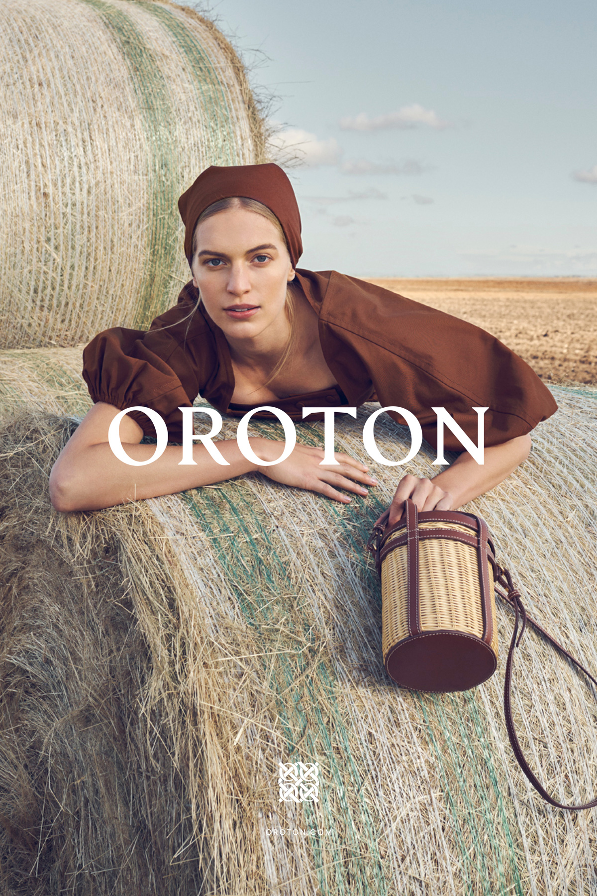 Woman in brown headscarf and puff-sleeve top laying on hay bales with designer handbag in outdoor rural setting under blue sky.