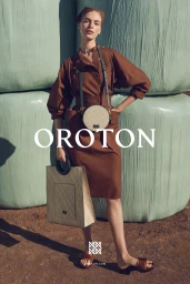 Vanessa Axente in brown dress and headscarf holding woven handbag standing before large pale green hay bales