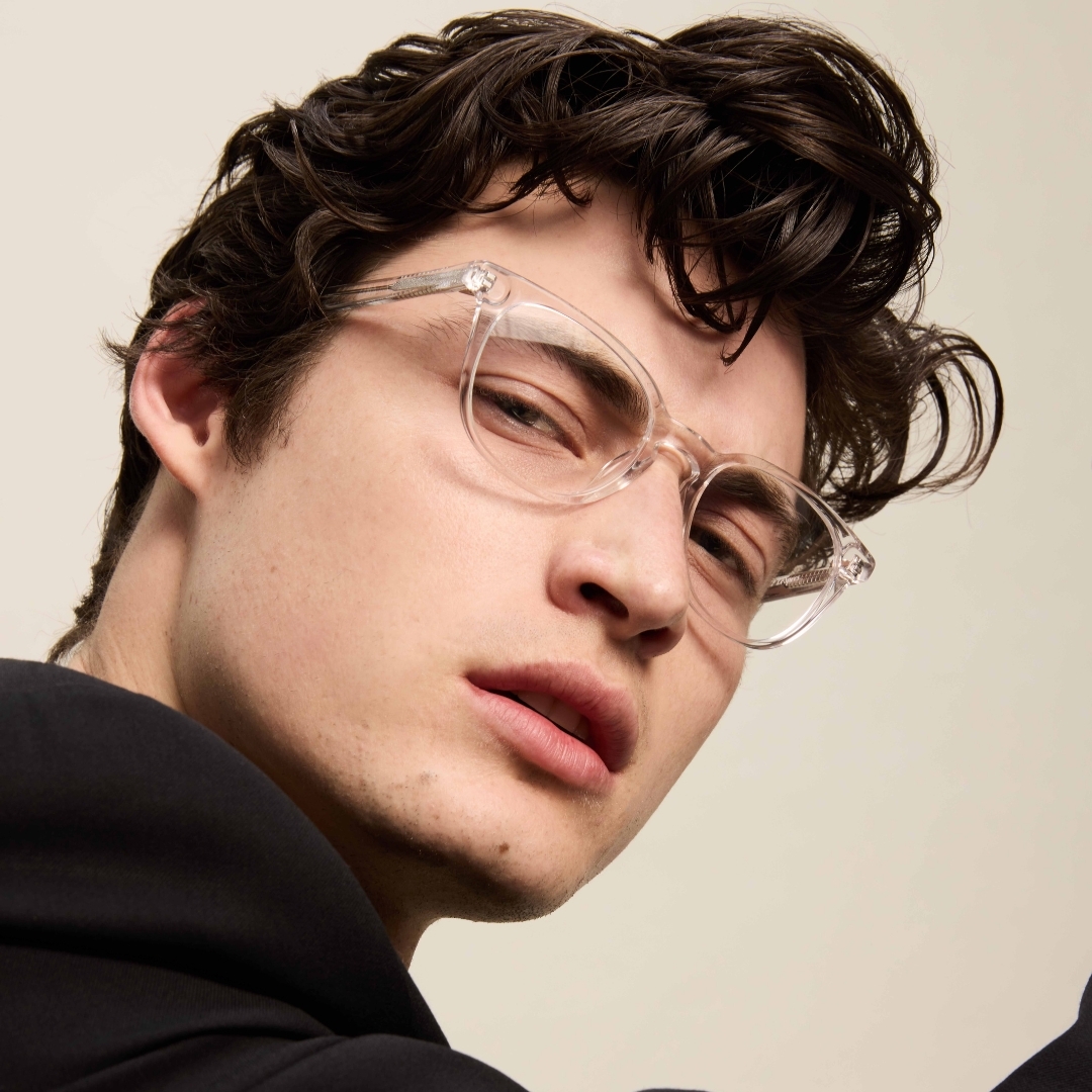 Close-up shot of a young adult male wearing transparent eyeglasses in a casual style, with a neutral expression against a plain background.