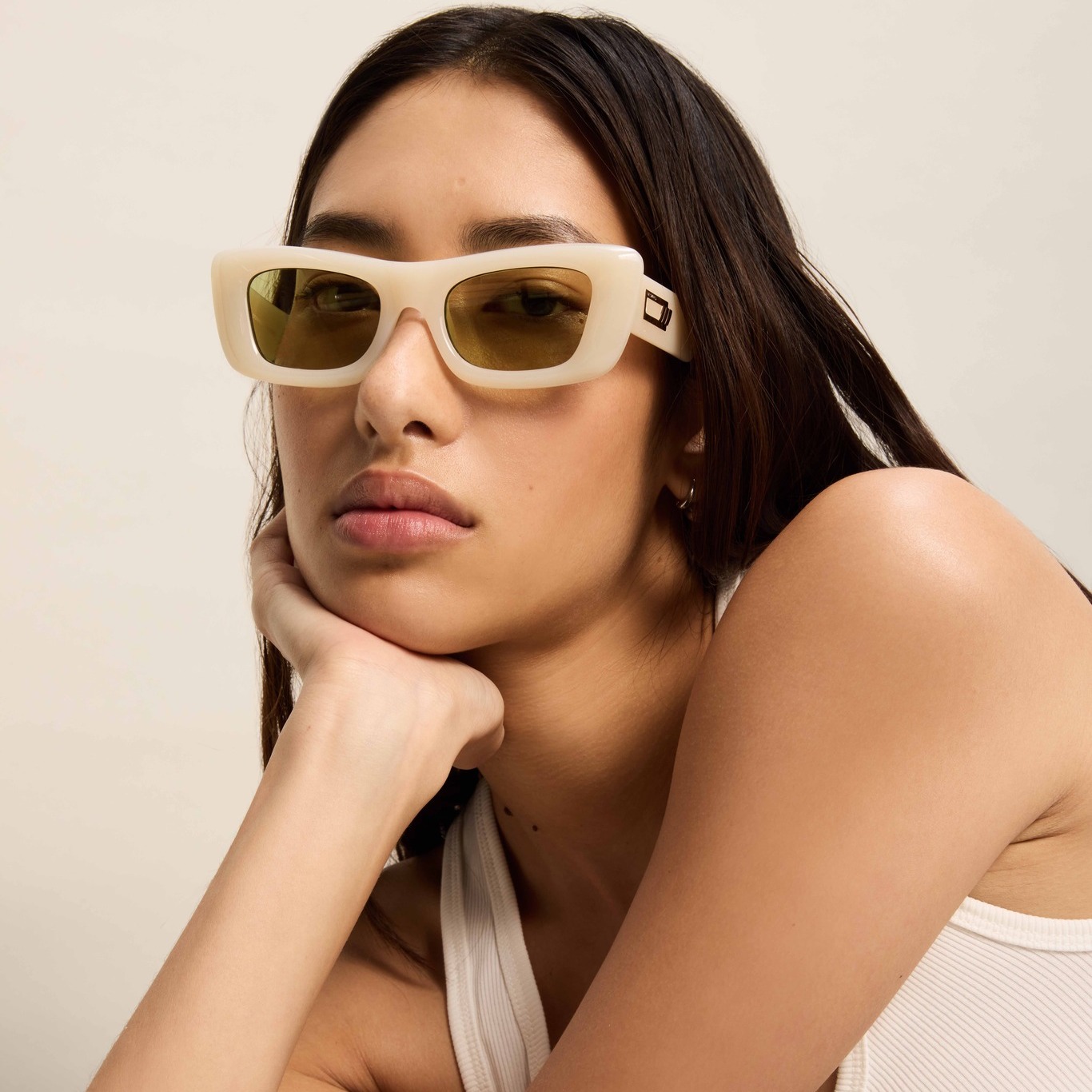 Brunette woman with tanned skin wearing white-rimmed, square sunglasses and a white tank top against a neutral background.