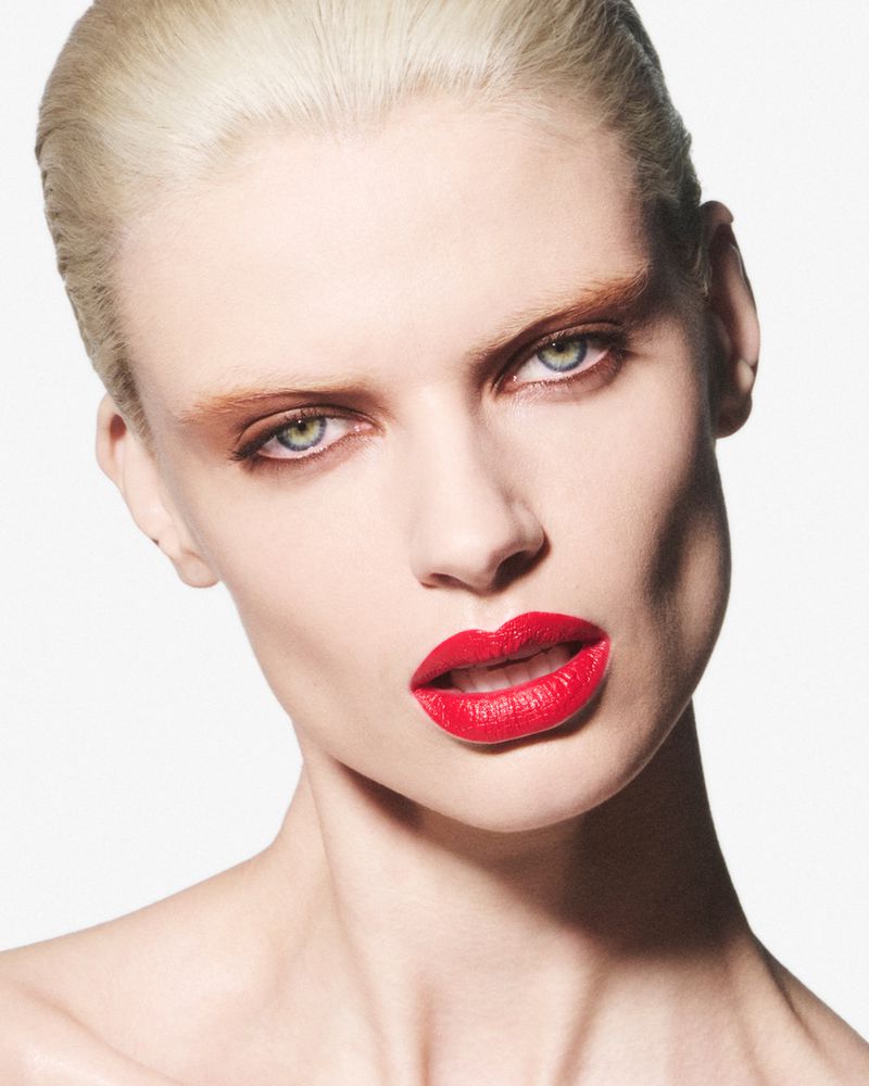 Close-up beauty portrait of a young adult Caucasian woman with slicked-back blonde hair and red lipstick against a white backdrop.