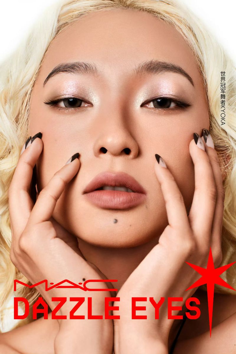 Beauty shot of a woman with blond curly hair, showcasing makeup and nail art, posing indoors against a white background.