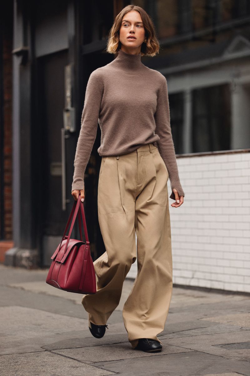 Model in brown turtleneck and beige trousers holding a burgundy leather handbag walking on a city street.