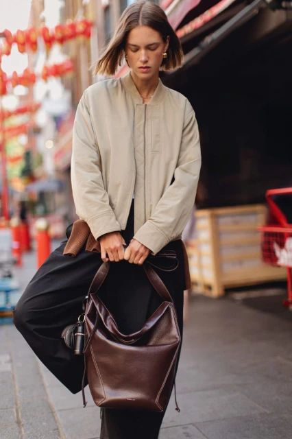 Woman in jacket and wide-leg pants carrying a large brown hobo bag, posing on a city street with red lanterns in the background.