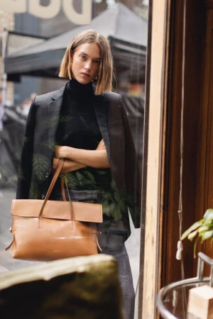 Fashion shot of a woman in black top and blazer, carrying a tan leather satchel, standing near a window with a blurred urban view.