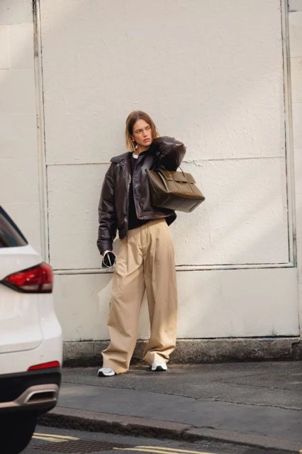 Street-style fashion shoot featuring a woman in a leather jacket, oversized pants, sneakers, carrying an olive green handbag outdoors.
