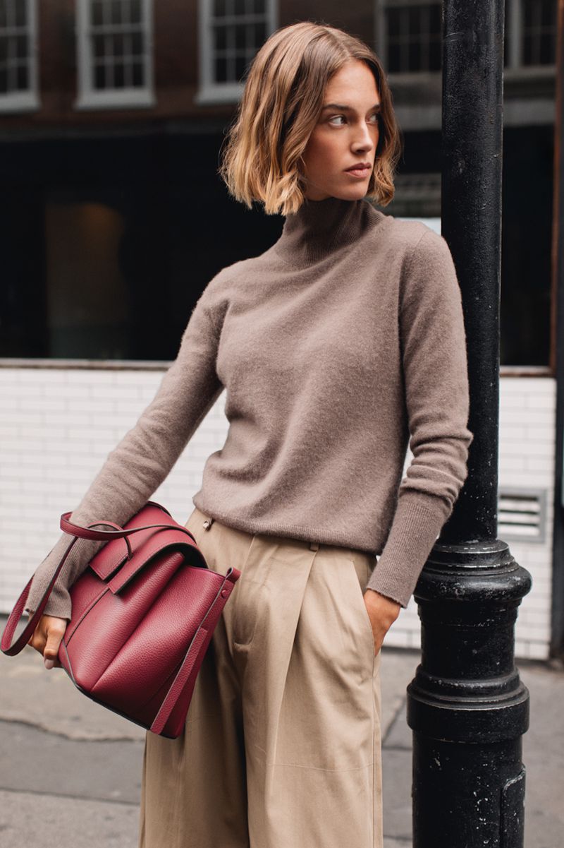 Adult woman poses in beige sweater and tan pants, holding a red leather tote bag in an urban setting with a street lamp.