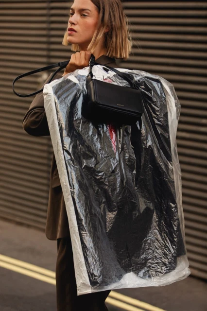 Woman carrying a garment bag with a black purse in an urban setting. She has short hair and is wearing a brown blazer.