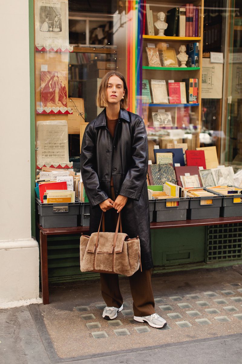 Woman in a black trench coat carrying a large brown suede tote bag standing in front of a book shop with books on display.
