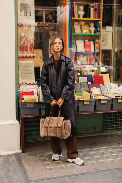 Woman in a black trench coat carrying a large brown suede tote bag standing in front of a book shop with books on display.