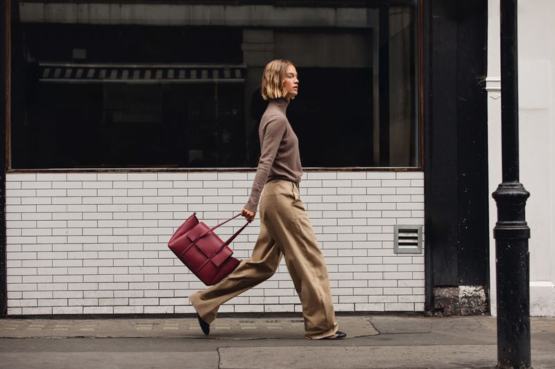 Model with blonde bob wearing a brown turtleneck and wide trousers walking past a tiled storefront holding a burgundy handbag
