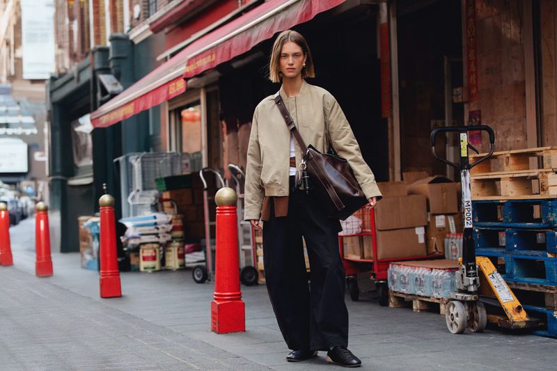 A young woman walks on a sidewalk in an urban setting, wearing a tan bomber jacket, dark pants, and carrying a large leather bag.
