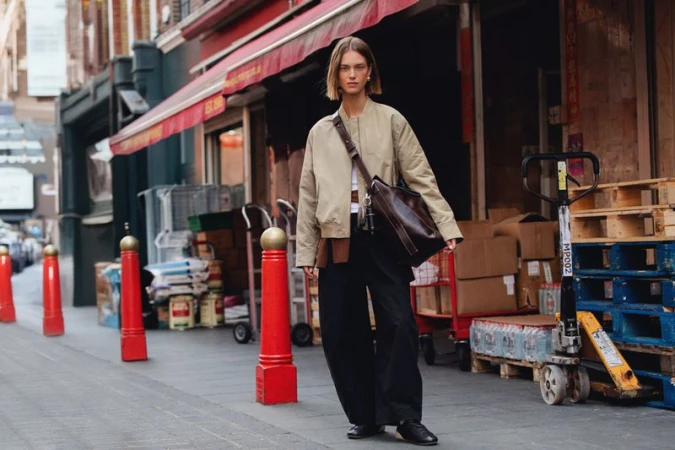 A young woman walks on a sidewalk in an urban setting, wearing a tan bomber jacket, dark pants, and carrying a large leather bag.