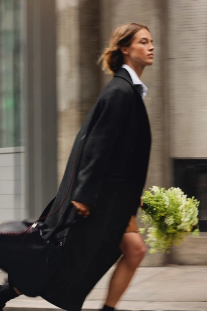 Fashionable woman in motion wearing a stylish black coat and carrying green flowers, walking on an urban city street with blurred motion.