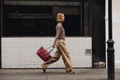 Model with blonde bob wearing a brown turtleneck and wide trousers walking past a tiled storefront holding a burgundy handbag