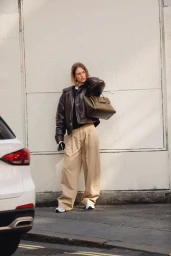 Model wearing dark leather jacket and beige trousers holding a leather handbag against a wall
