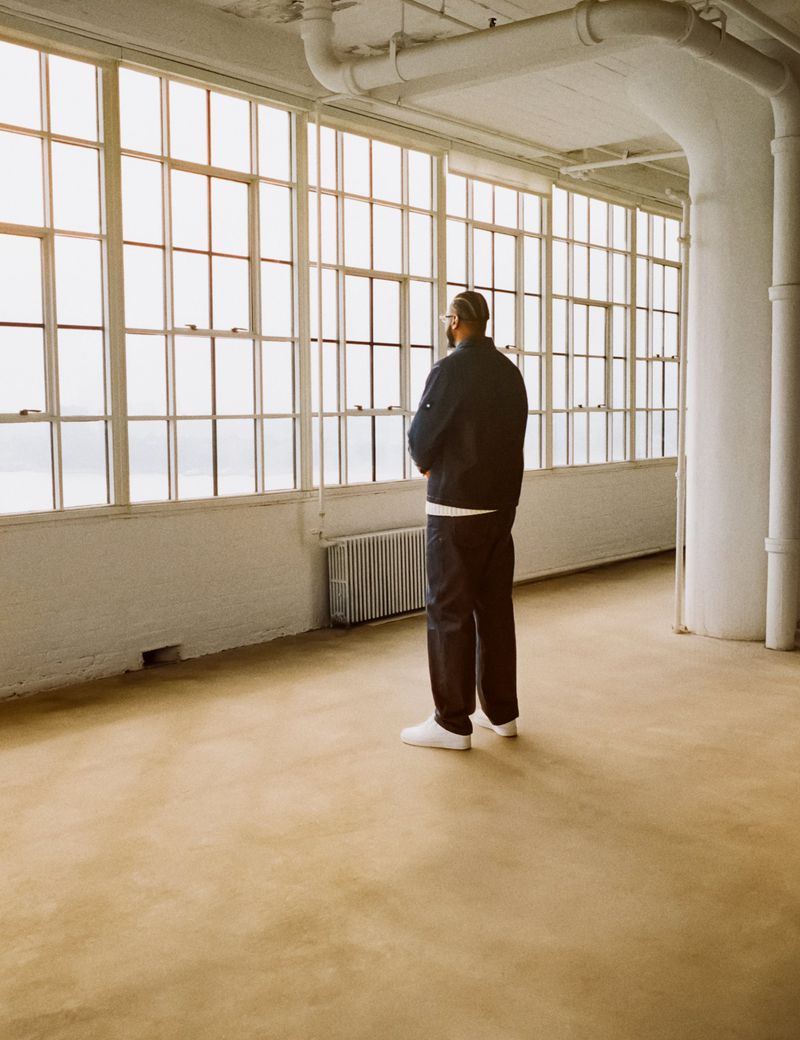 Adult male stands in sunlit loft apartment interior wearing dark blue jacket, pants, and white sneakers, looking out large window.