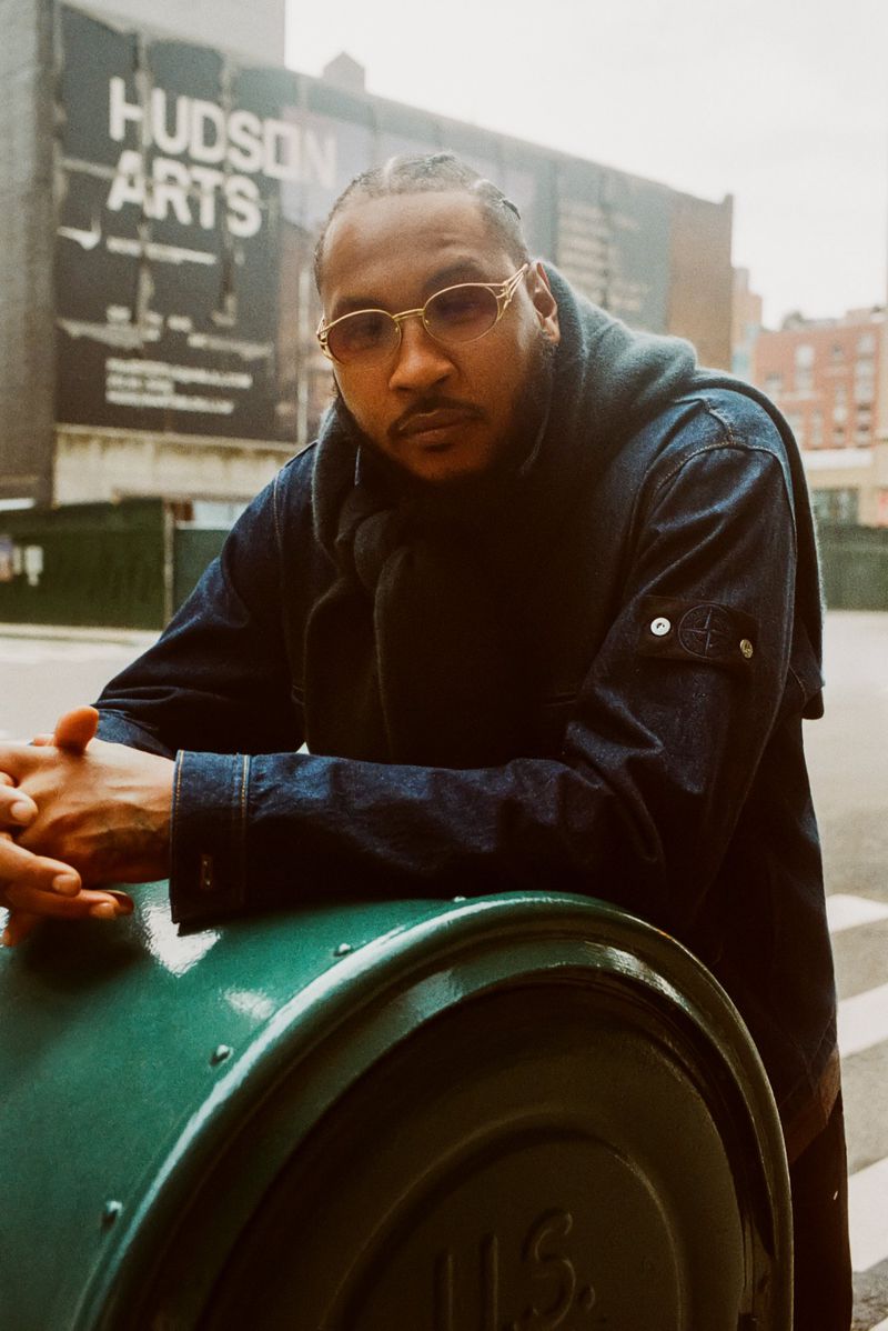 Adult man poses against a green mailbox on a city street, wearing gold glasses and a blue jean jacket with a poster in the background.