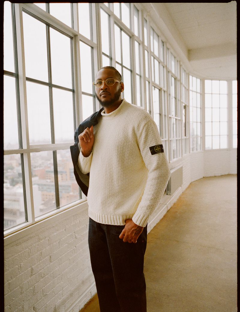 Portrait of a stylish man in a cream sweater and black pants standing near large windows with a jacket over his shoulder.